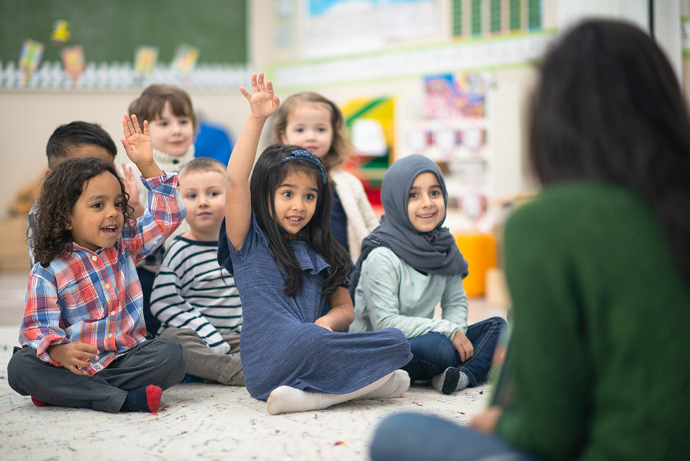 A multi-ethnic group of preschool kids is indoors in their classroom. They are sitting on the floor and raising their hand to answer a question posed by their teacher while reading a story. They are all smiling.